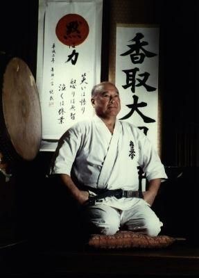 Masutatsu Oyama w białym karategi siedzi na klęczkach na tle kaligrafii